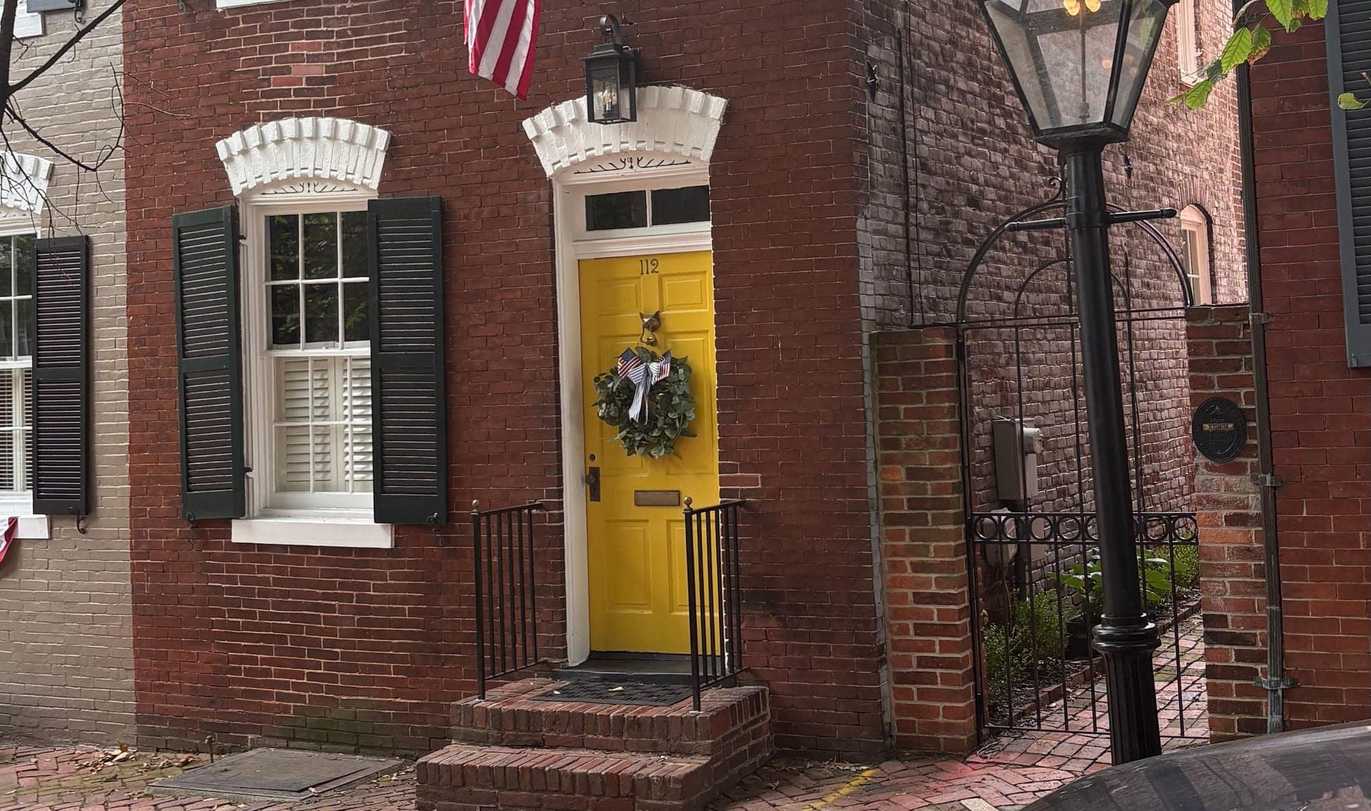 A bright yellow front door with a wreath on a red-brick Old Town Alexandria rowhouse, beside a gas lamp