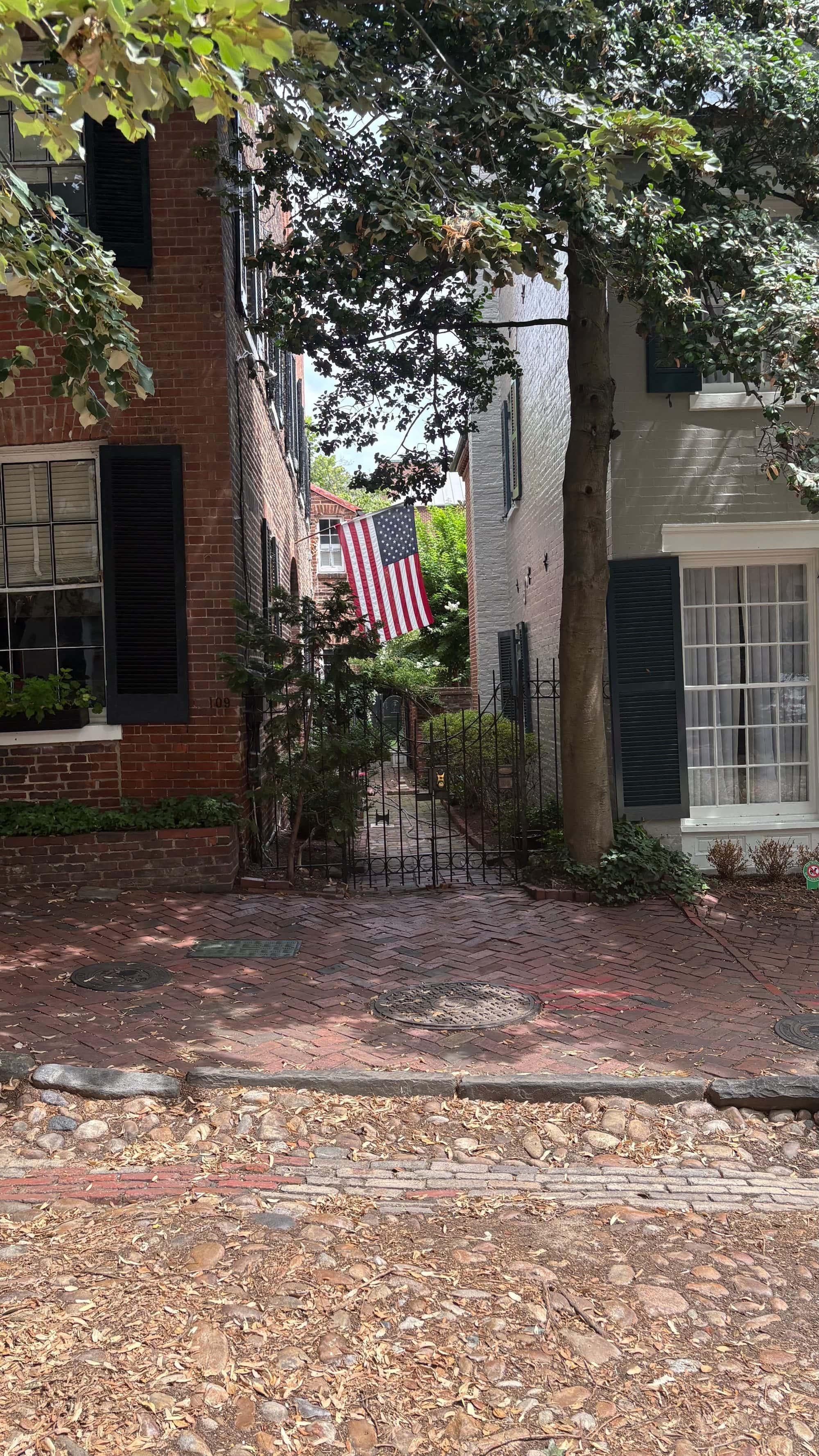 A quiet Old Town Alexandria brick alley leading to a wrought-iron garden gate, an American flag hanging between the rowhouses